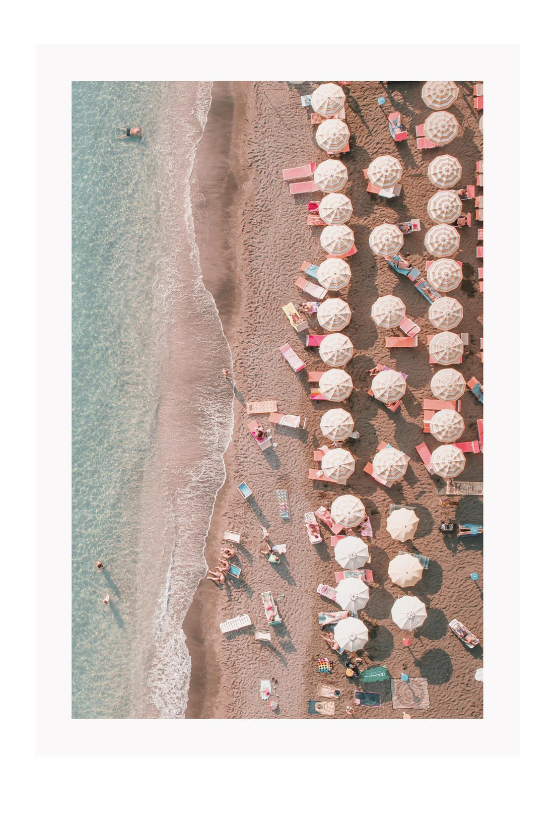 Coastal style photography beach print with pink umbrellas and chairs on the sand in front of the water from a birds eye view.